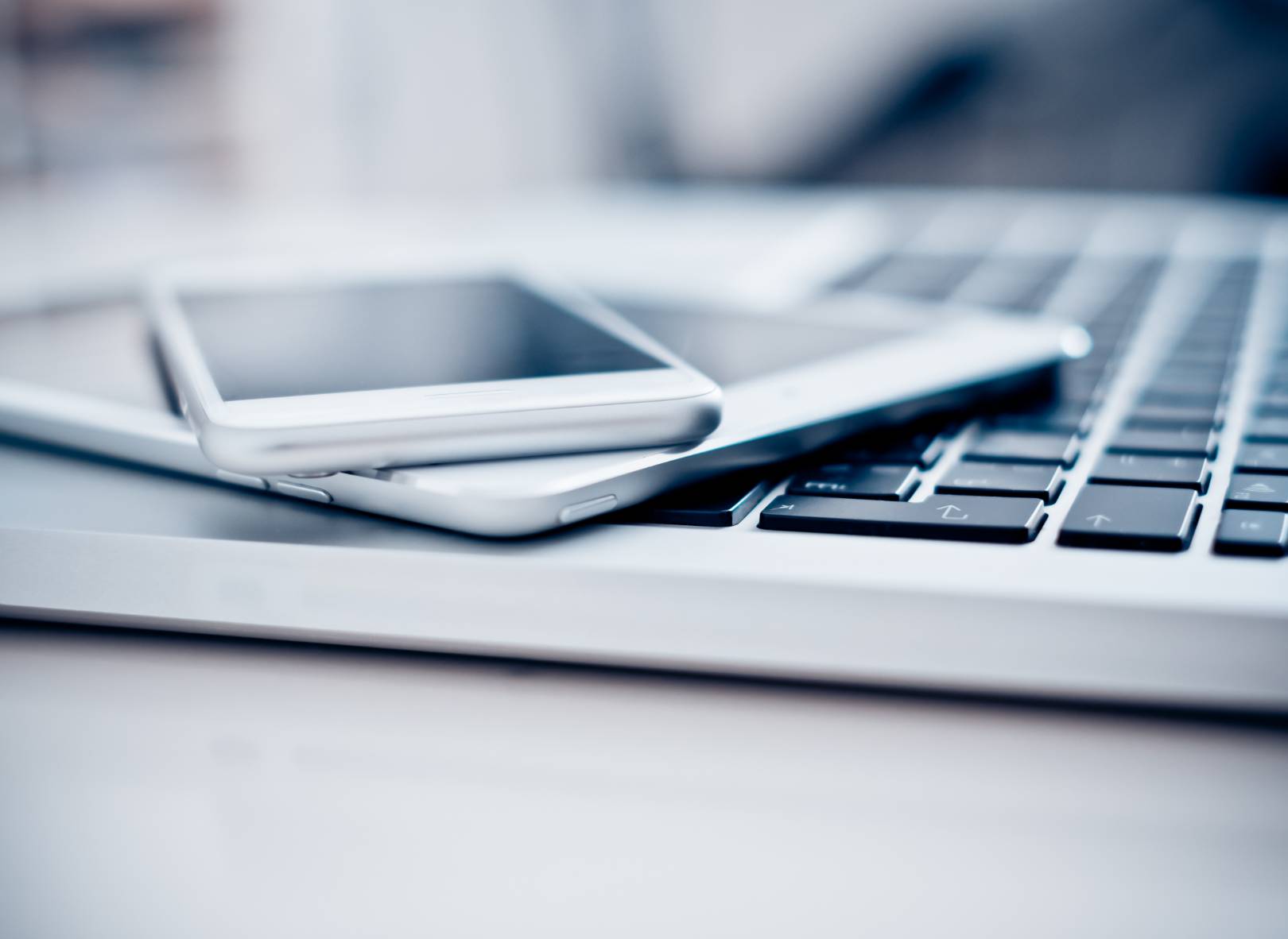 Two smartphones stacked on top of each other, resting on the keyboard of an open laptop. The devices are all silver or white and the image is softly focused, highlighting a modern tech workspace.