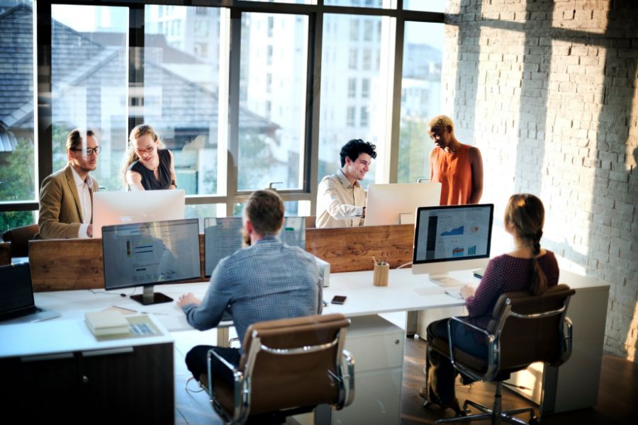 Six people work together in a modern, sunlit office with large windows. Some are seated at desks with computers displaying charts, while others stand and discuss near the monitors. The atmosphere appears collaborative and focused.