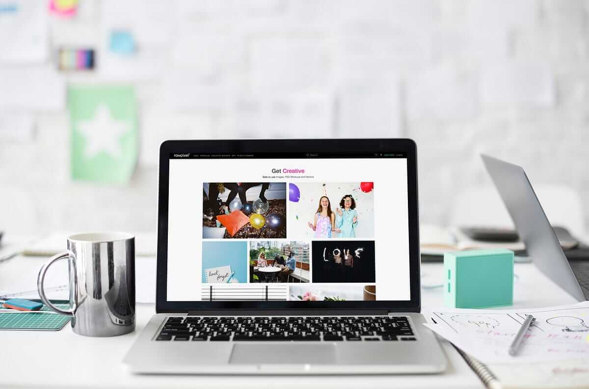 A laptop on a white desk displays a creative inspiration website. Next to it are a silver mug, a smartphone, a blue box, papers, and office supplies. The background shows a blurred wall with notes and photos.