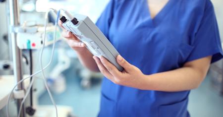 Healthcare professional in blue scrubs using a digital interface to monitor and manage medical devices in a hospital setting.