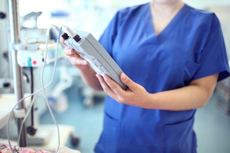 Healthcare professional in blue scrubs using a digital interface to monitor and manage medical devices in a hospital setting.