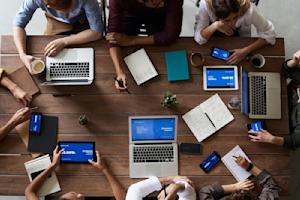 A group of employees sitting around a table and using several devices including laptops, tablets, and smartphones.