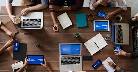 A group of employees sitting around a table and using several devices including laptops, tablets, and smartphones.