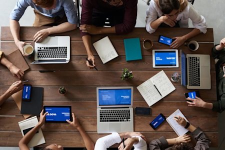 A group of employees sitting around a table and using several devices including laptops, tablets, and smartphones.