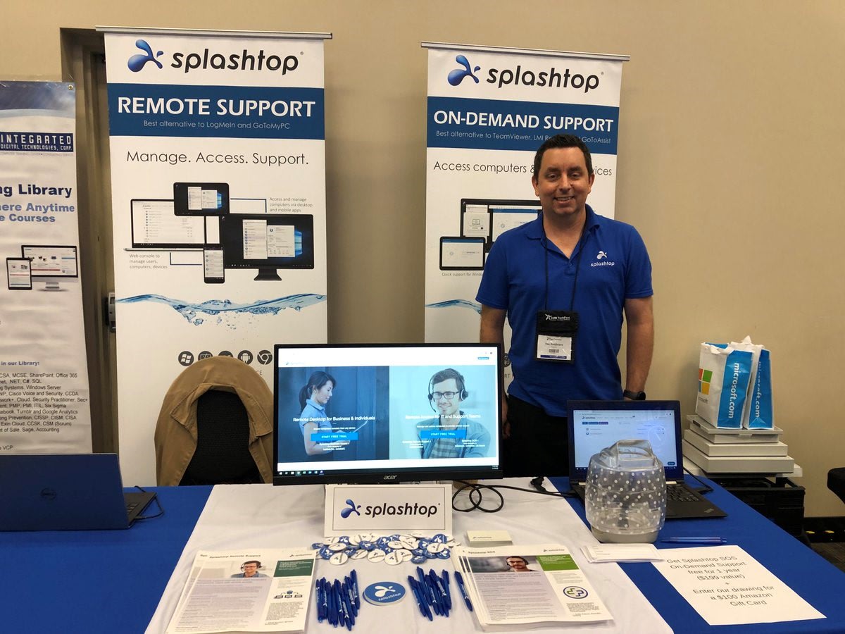 A man stands at a Splashtop booth featuring banners for Remote Support and On-Demand Support. The table displays a monitor, brochures, pens, buttons, and a raffle bowl at a conference or expo.