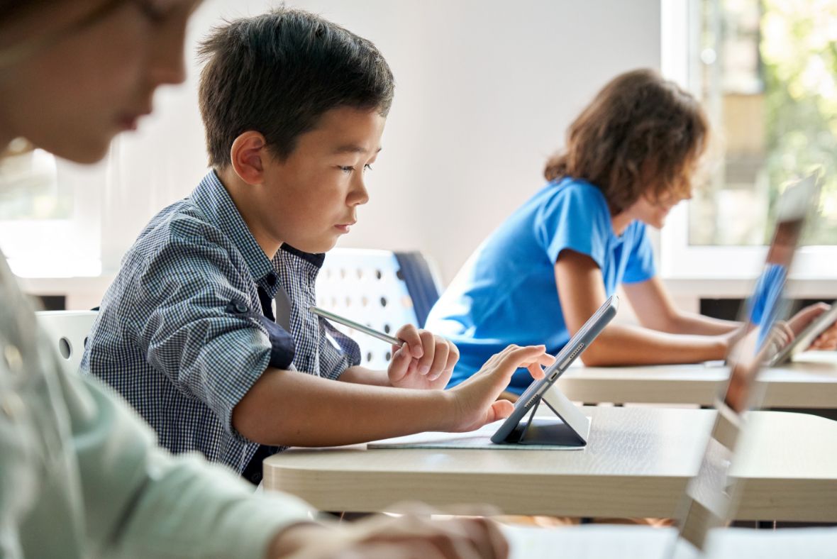 Students in a bright classroom working on tablets, with a boy in checkered shirt focused on his device.