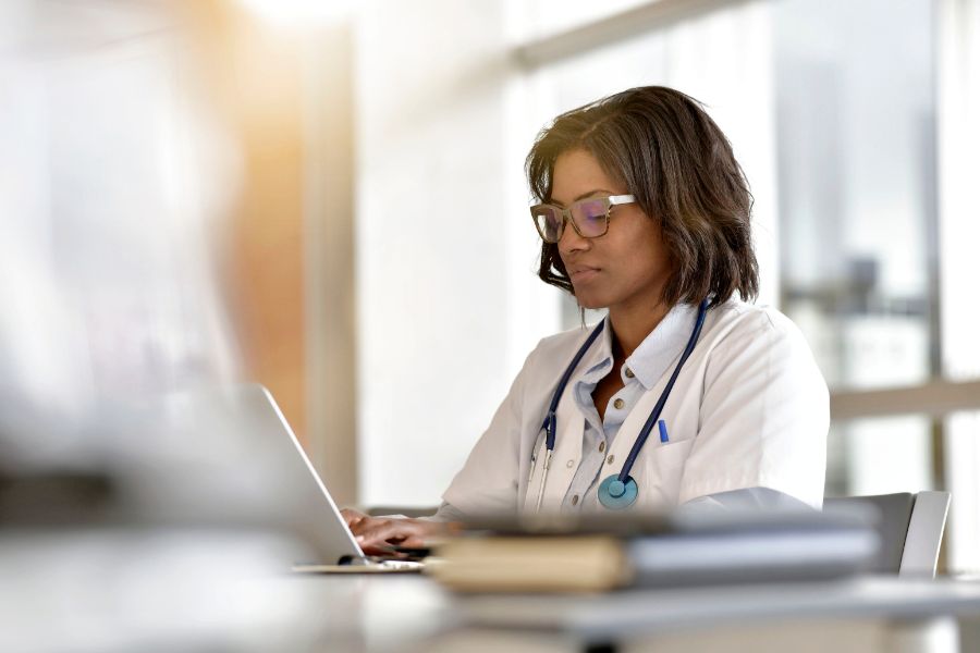 A doctor wearing a white coat and stethoscope sits at a desk, focused on working on a laptop in a bright, modern office setting.