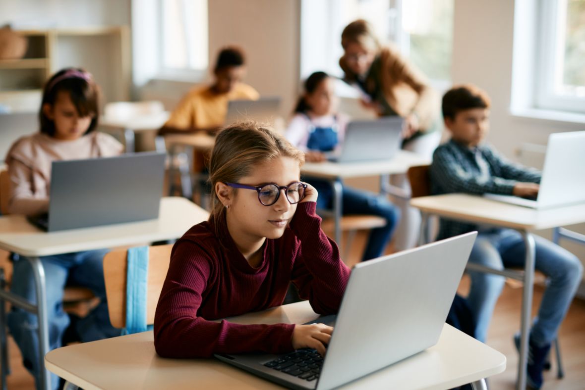A young girl wearing glasses sits at a desk using a laptop in a classroom, with other students also working on laptops and a teacher assisting in the background.