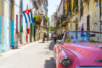 A pink convertible is photographed parked in an alleyway located somewhere in an urbanised area of Cuba. The Cuban flag is visible hanging off a flagpole protruding from a building in the midground of the photograph