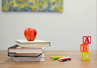 A red apple sits atop four books stacked on a teacher-s desk next to a selection of coloured pencil and a stack of lettered wooden playing blocks