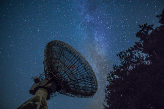 An astronomical radar dish points towards the night sky with milky way gas formations enhanced in the background