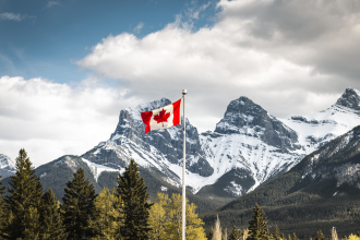 A Canadian flag flies high atop a flagpole carried by the wind in the foreground of an alpine horizon beneath a cloudy but pleasant sky