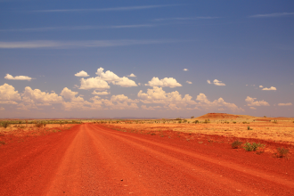 A pindan dirt road stretches into the outback horizon like a giant petroglyph beneath a blue sky; no trees are visible for miles aroind