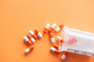 A generic image of prescription medication spilling out of a canister designed to hold it against an orange tabletop background