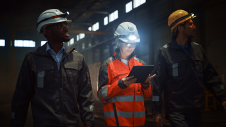 Three workers in a mining environment travel through a facility; a warden in between two colleagues inspects information on a touhscreen tablet; her helmet light illuminates a safety vest in relative darkness