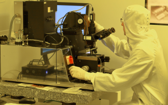 Engineer in tech cleanroom wearing protective clothing inspects a product through a microscope 