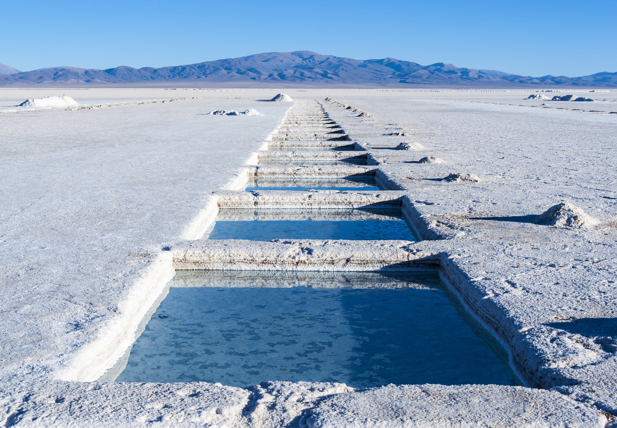 A row of lithium brine ponds extend into the distance in a straight line from the photographer; mountains cover the horizon in the distance. 