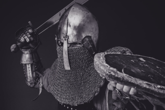 A black and white image of a model dressed in replica knight-s armour posing in a combative position against a black background