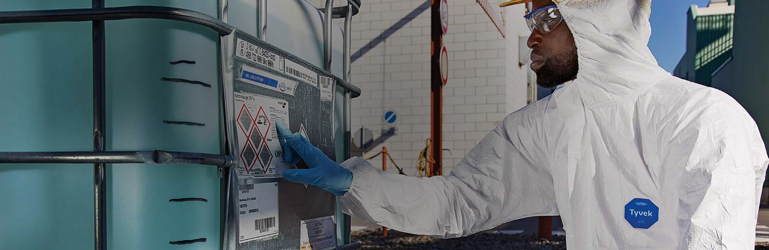 Worker outside facility wearing DuPont Tyvek 500 coverall checking hazard label on large container.