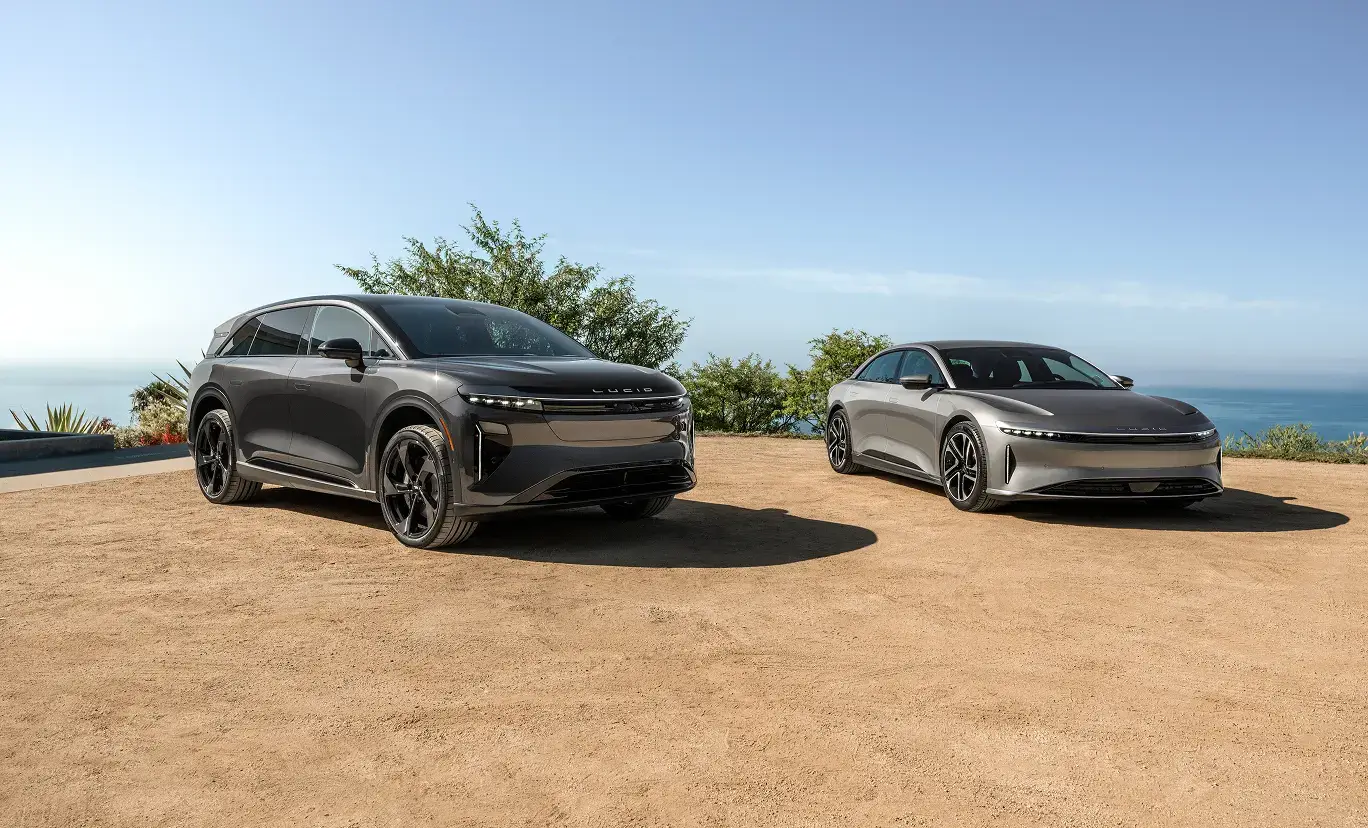 Two Lucid electric vehicles—a dark gray SUV and a silver sedan—parked on a sandy overlook with ocean views.