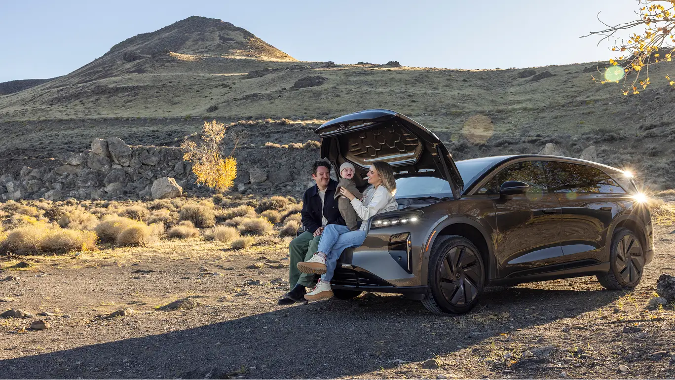 Lucid Gravity SUV parked in desert landscape with mountain backdrop, open frunk, people sitting in frunk at sunset.