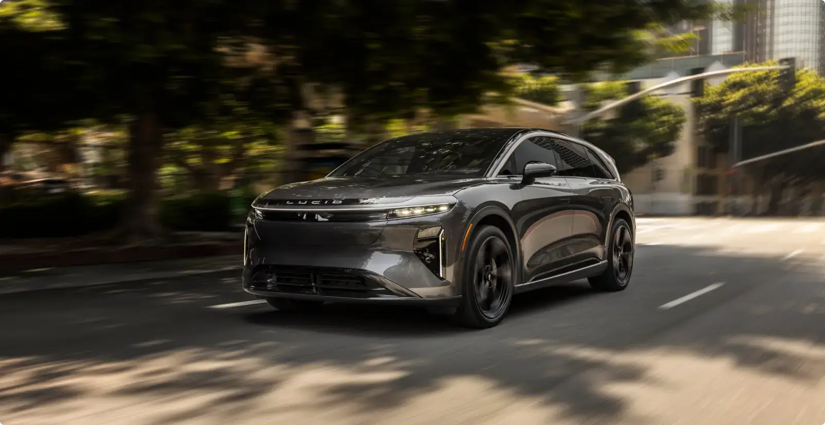 A gray Lucid electric SUV driving on a city street, with blurred trees and buildings in the background.