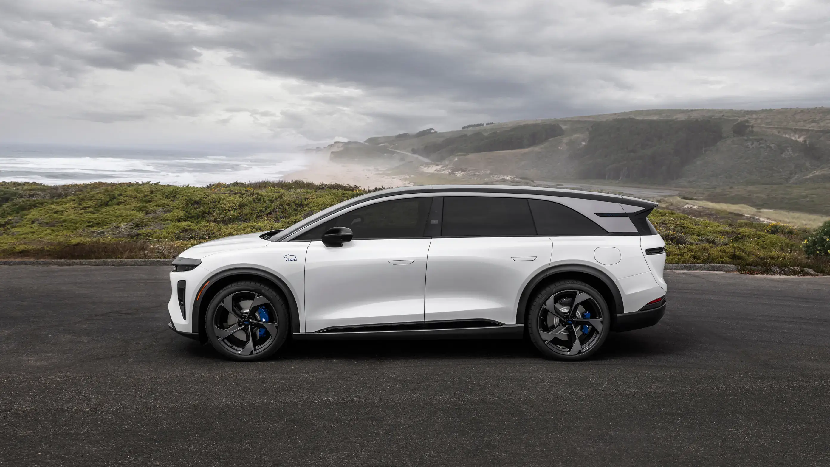 White electric SUV with black roof and blue brake calipers parked on coastal road with ocean cliffs and cloudy sky.