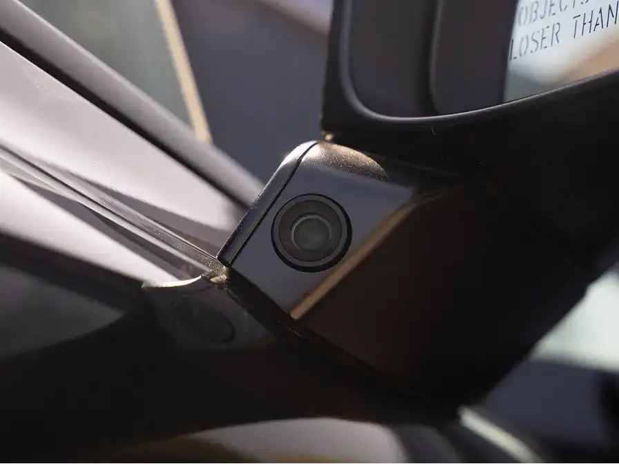 Close-up of a small black camera lens mounted on a car's side mirror housing, with a side mirror visible nearby.