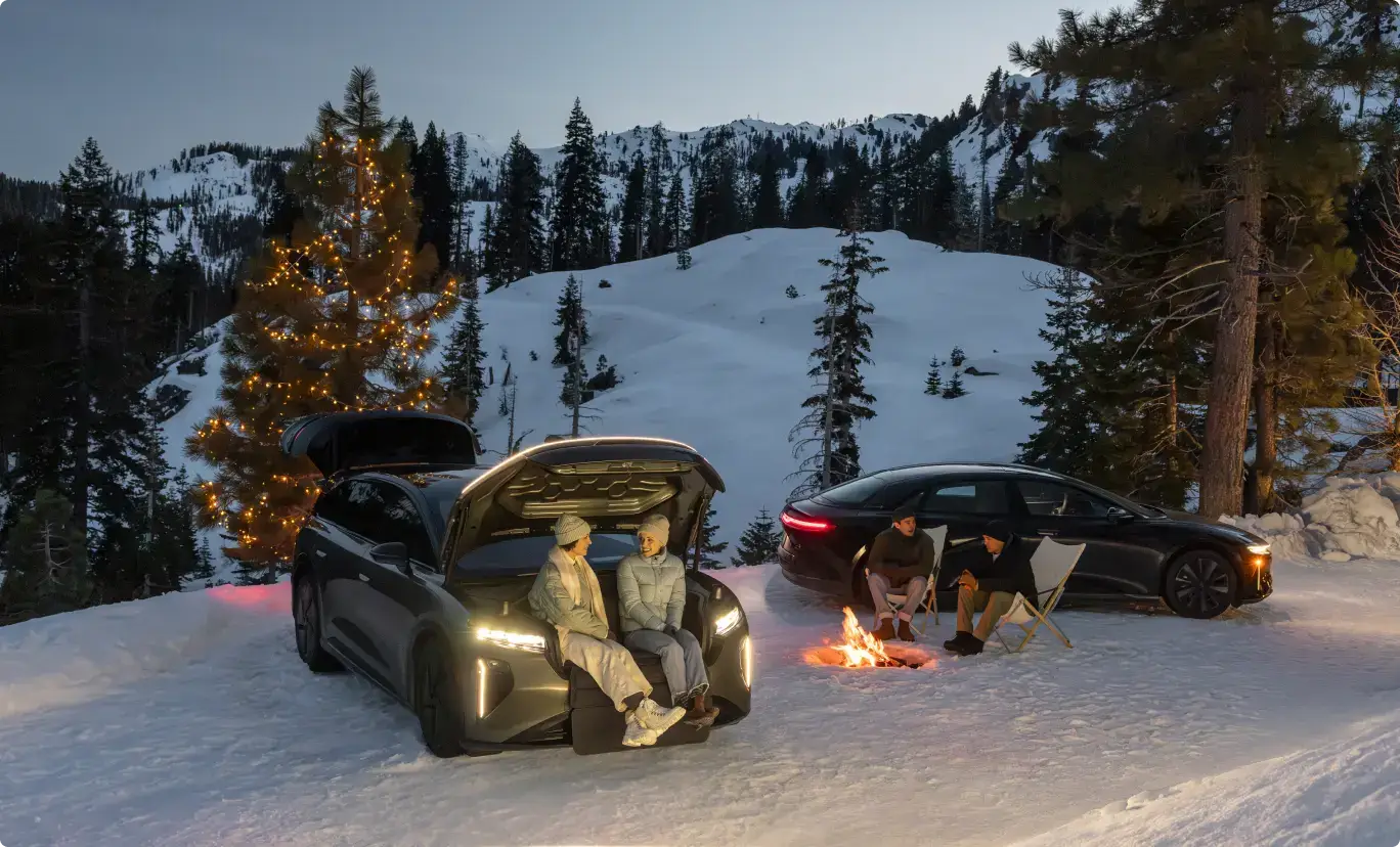 People enjoying a winter evening with two vehicles parked in snow, campfire, and a pine tree decorated with lights.