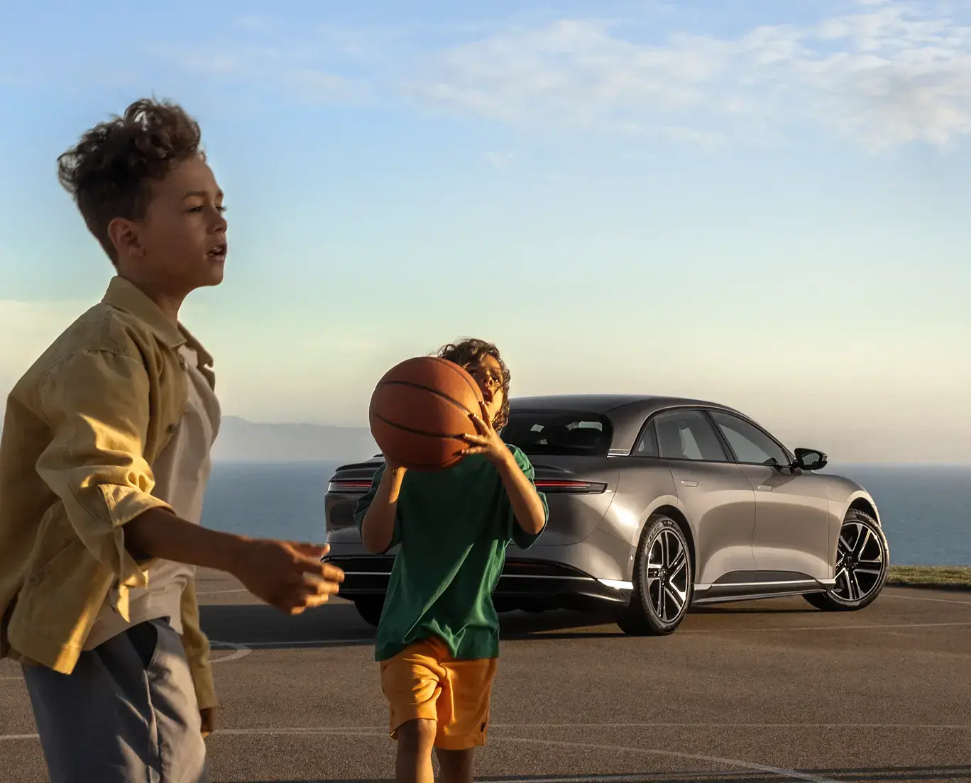 Two children playing basketball outdoors near a silver luxury sedan parked by a scenic coastal overlook.