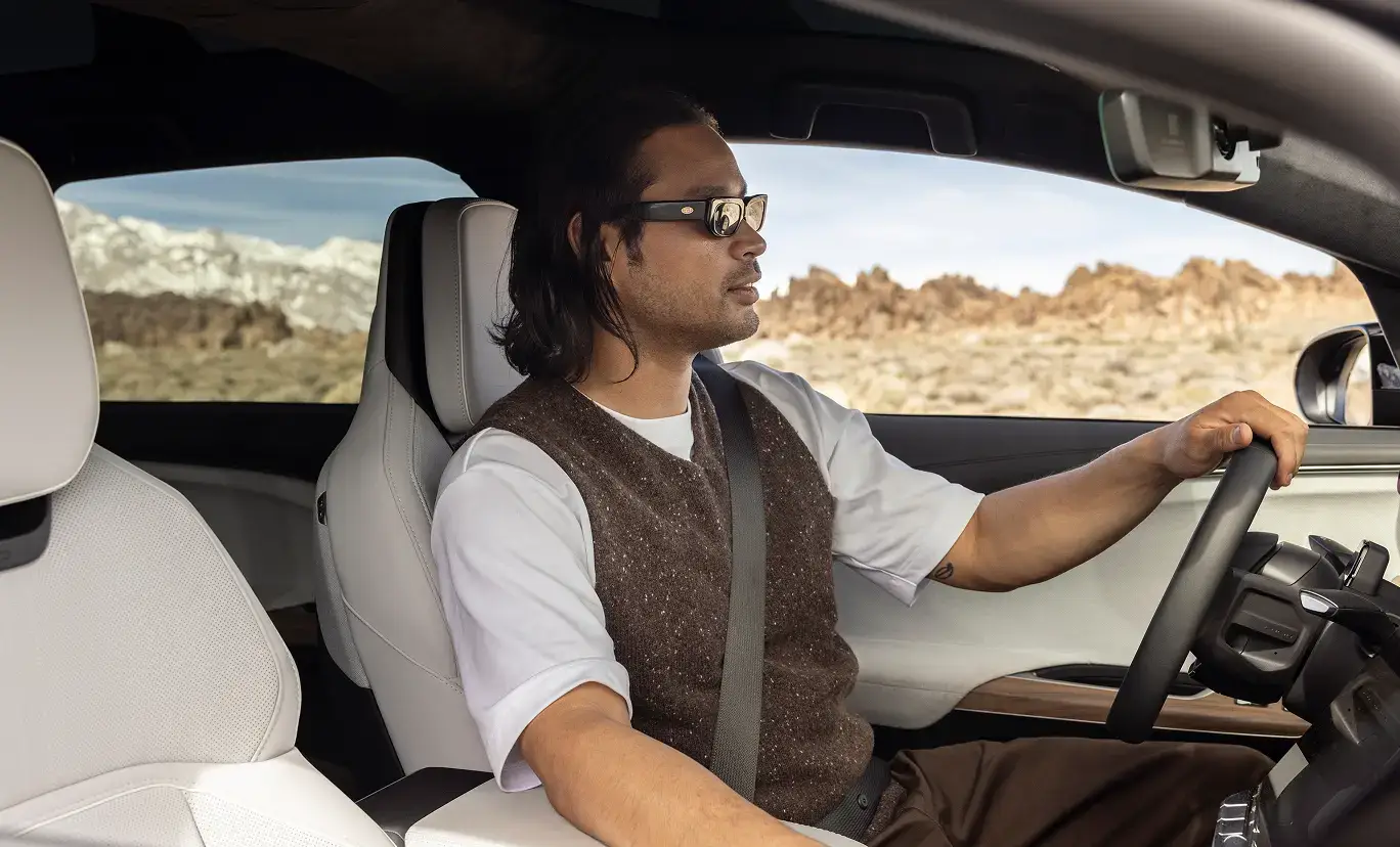 Man with sunglasses driving a car with light colored interior, rocky desert landscape visible through the windshield.
