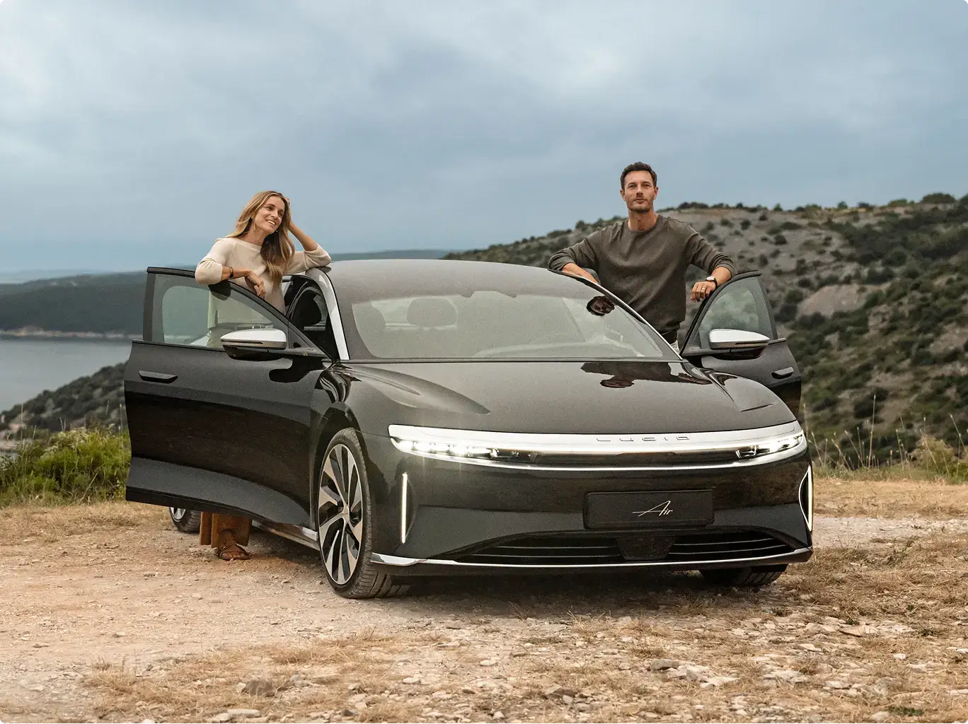 A man and woman stand beside a dark Lucid Air electric sedan outdoors, with hills and a lake in the background.