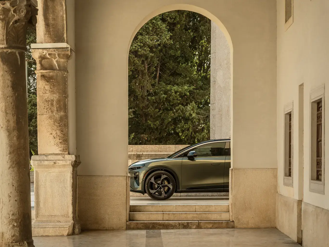 A green SUV parked outside, framed by a stone archway with columns, with lush green trees in the background.
