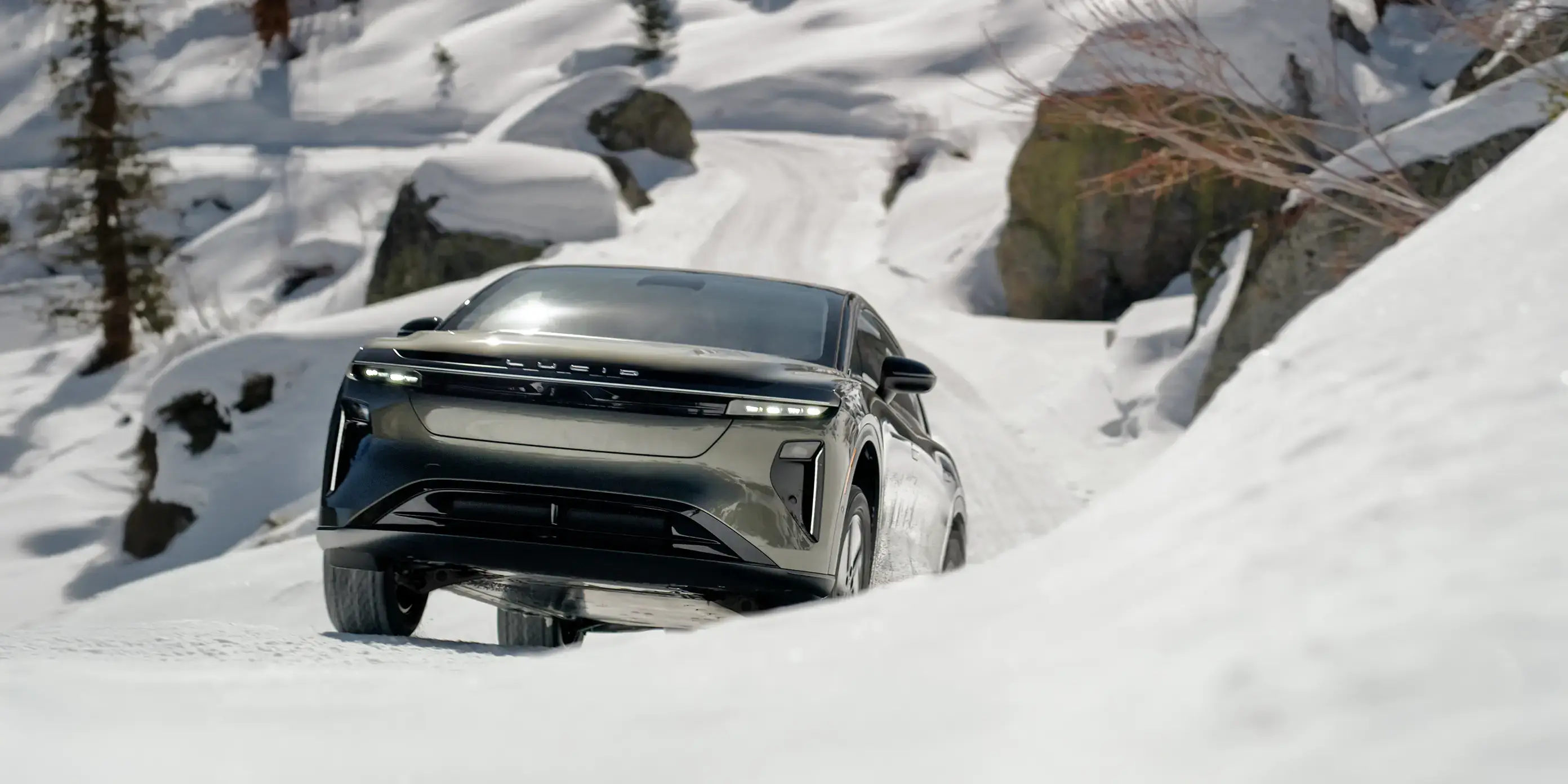 Modern Lucid Gravity vehicle driving through snowy mountain terrain with rocks and trees in background.

