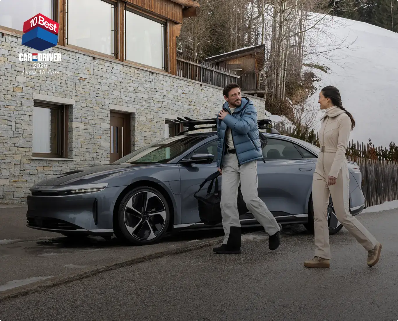 Two people in winter attire walking past a sleek gray car with ski rack outside a stone lodge, Car and Driver 10Best award shown.
