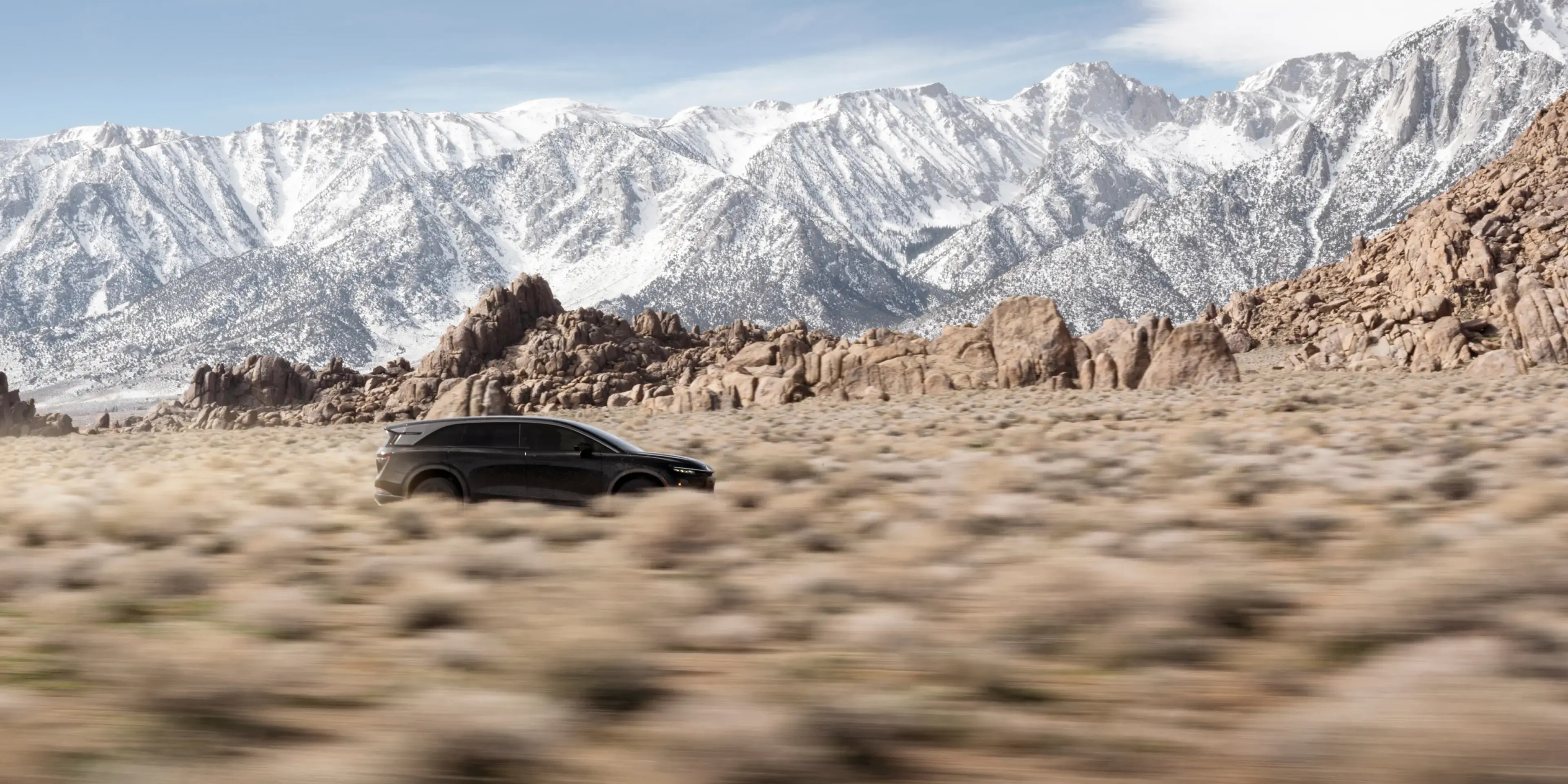 Lucid Gravity driving on desert road with snow-capped mountains and dramatic cloudy sky in background.