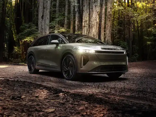 Aurora Green Metallic Lucid Gravity with illuminated headlights parked on a forest path among tall redwood trees at dusk.