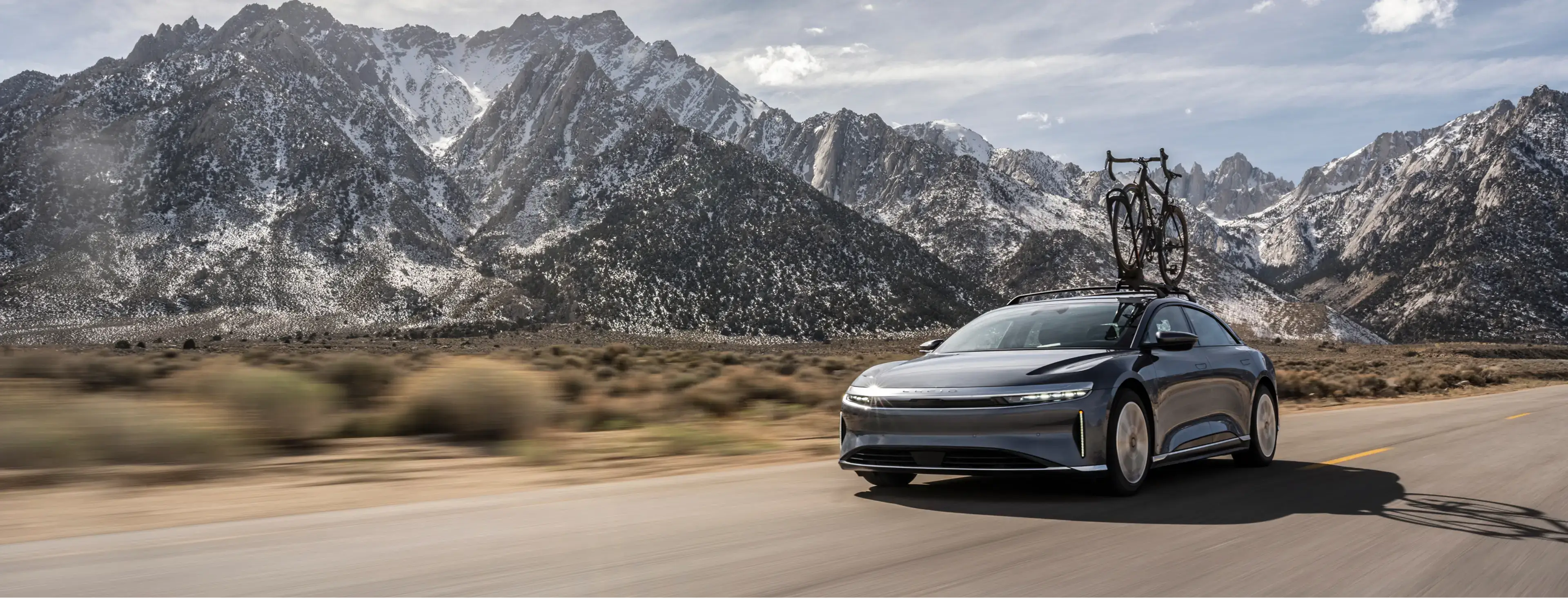 A Lucid Air electric sedan with a rooftop bike rack drives along a desert highway with snow-capped mountains behind.
