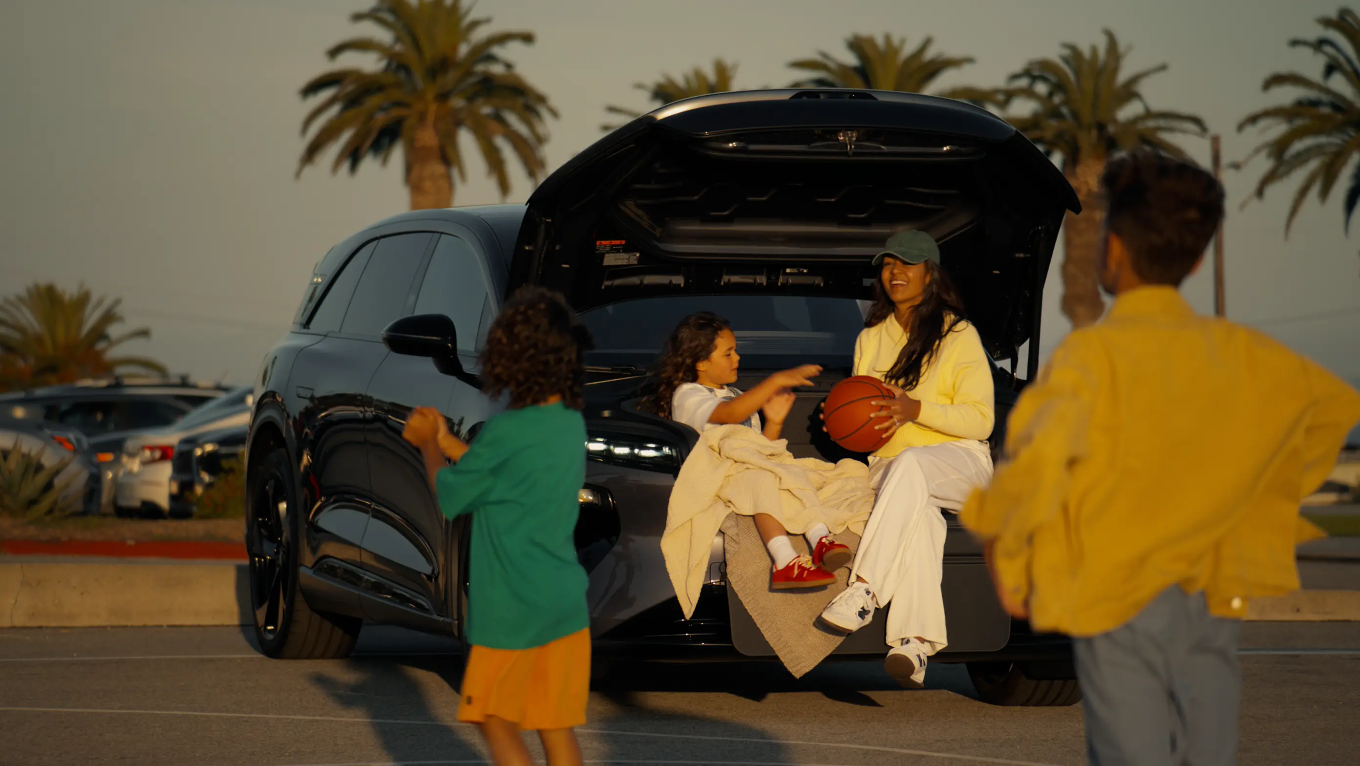 A woman holding a basketball sits in the open trunk of a Lucid SUV, smiling at children around her near palm trees.