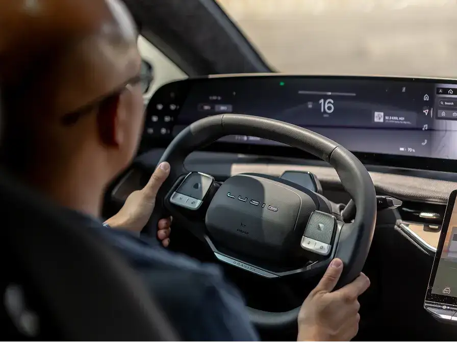 A driver grips a modern Lucid Gravity SUV steering wheel, with a large curved digital dashboard display visible ahead.