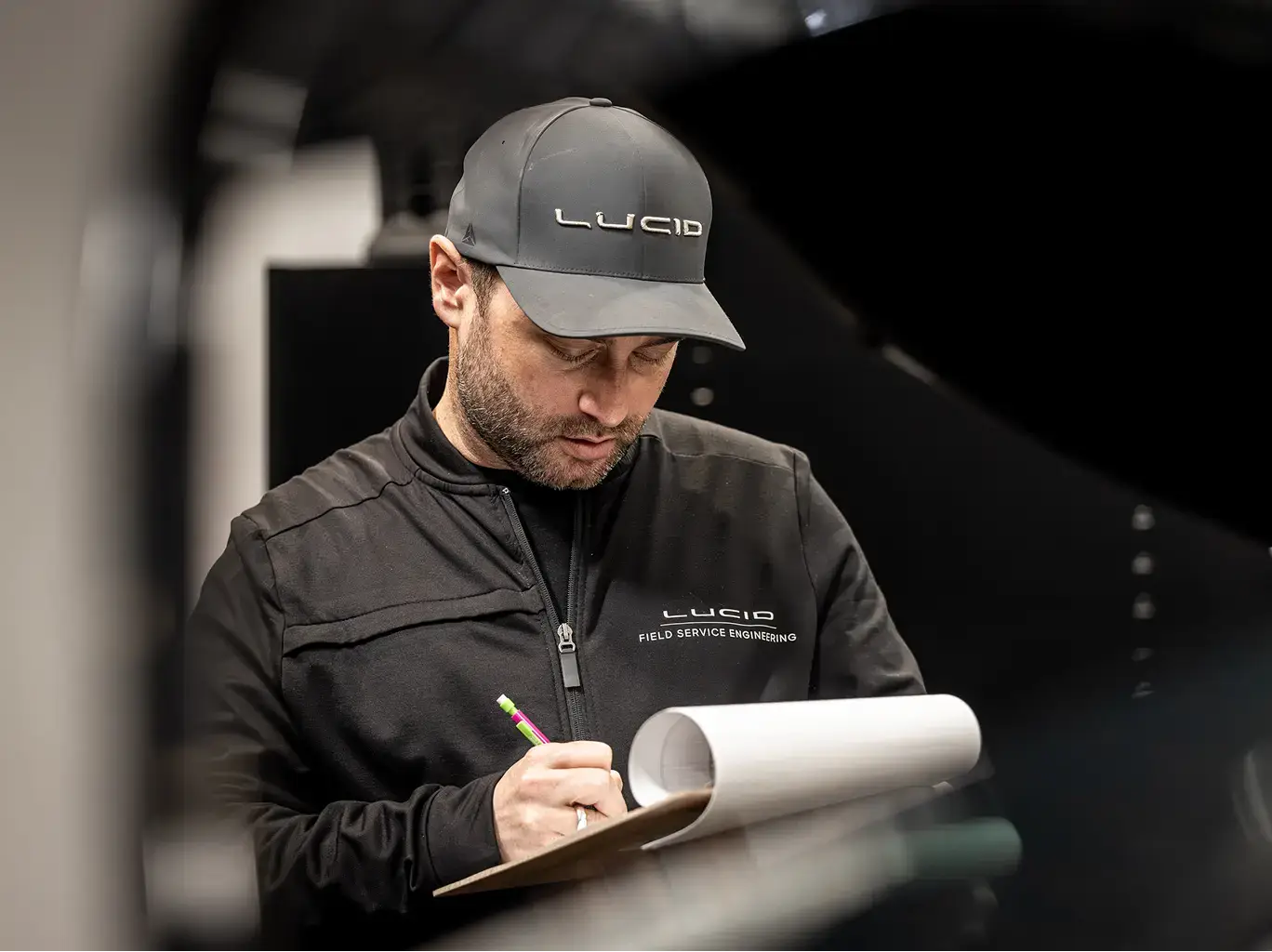 Lucid Field Service Engineering technician in black jacket and gray cap taking notes on clipboard with rolled documents.
