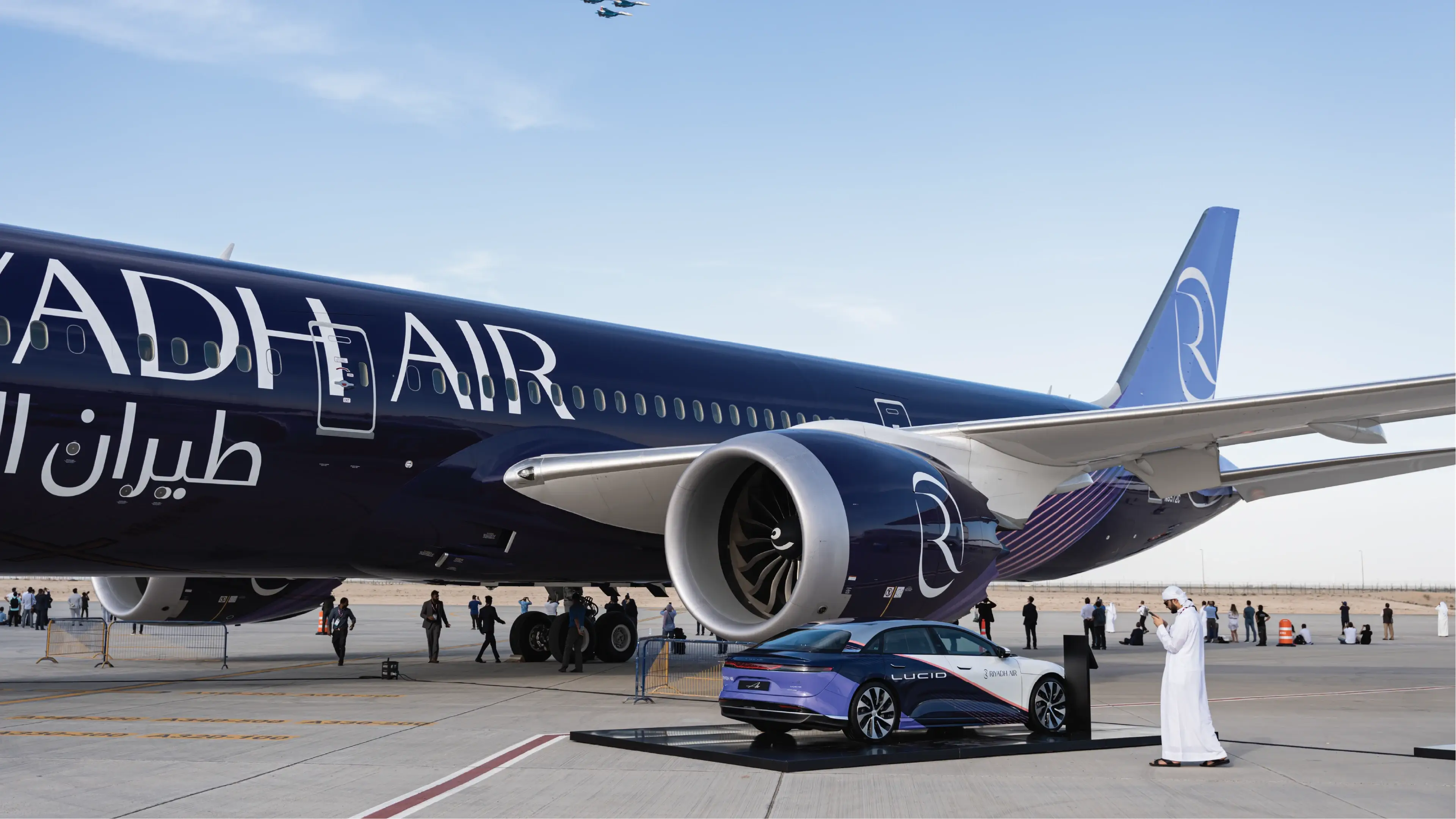 A large Riyadh Air airplane is parked on an airport tarmac beside a modern Lucid Air EV. The scene is calm, with clear skies and a few people nearby.