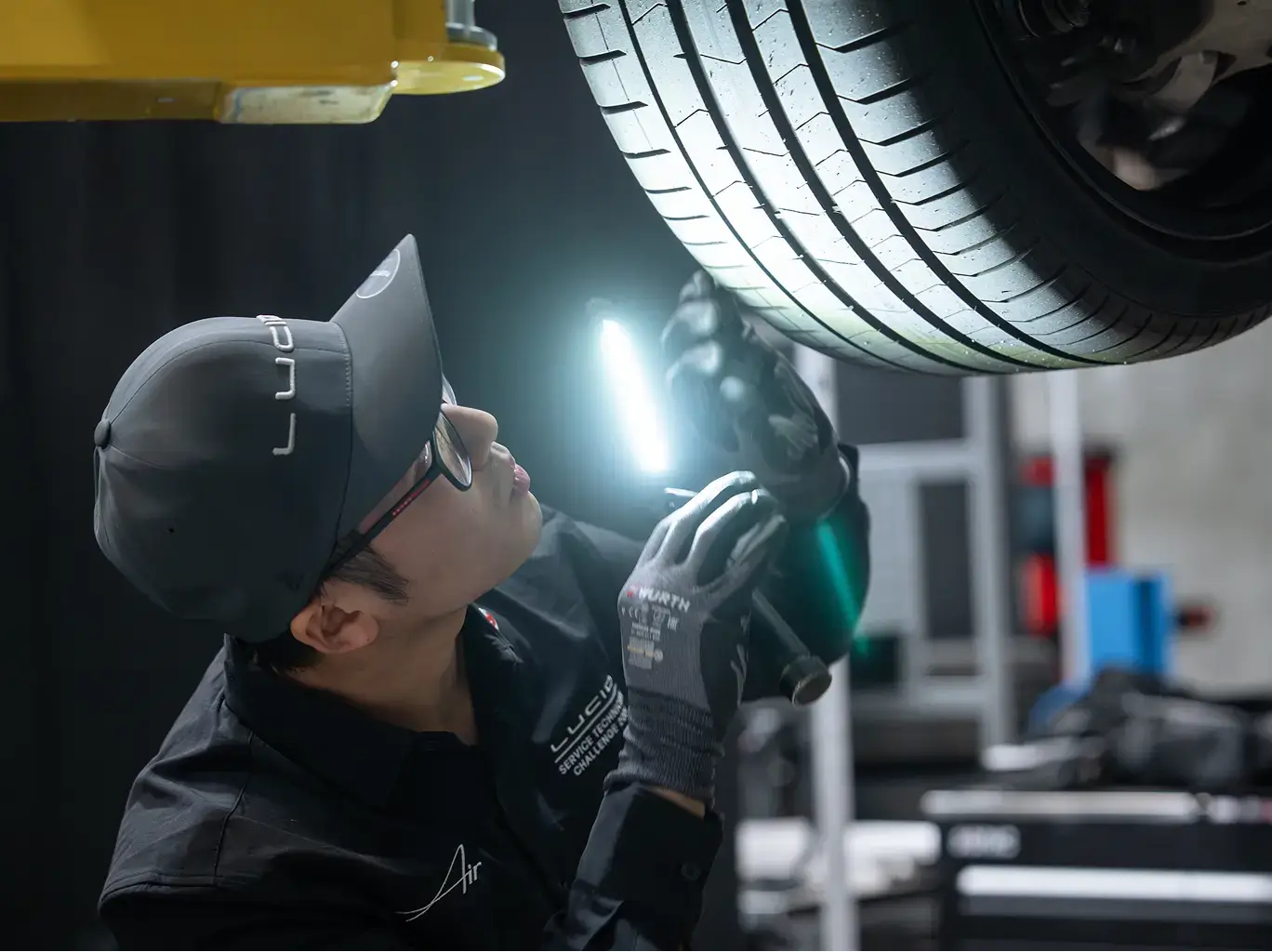 Mechanic in cap and gloves inspecting car tire tread with flashlight in auto repair garage