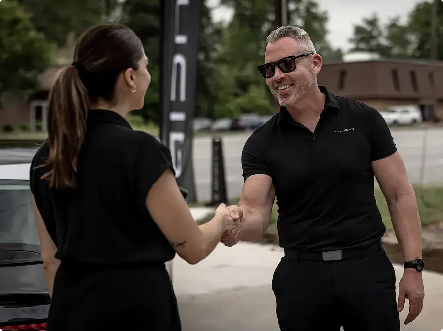A Lucid Motors employee in a black polo shakes hands with a woman outdoors near a car dealership.