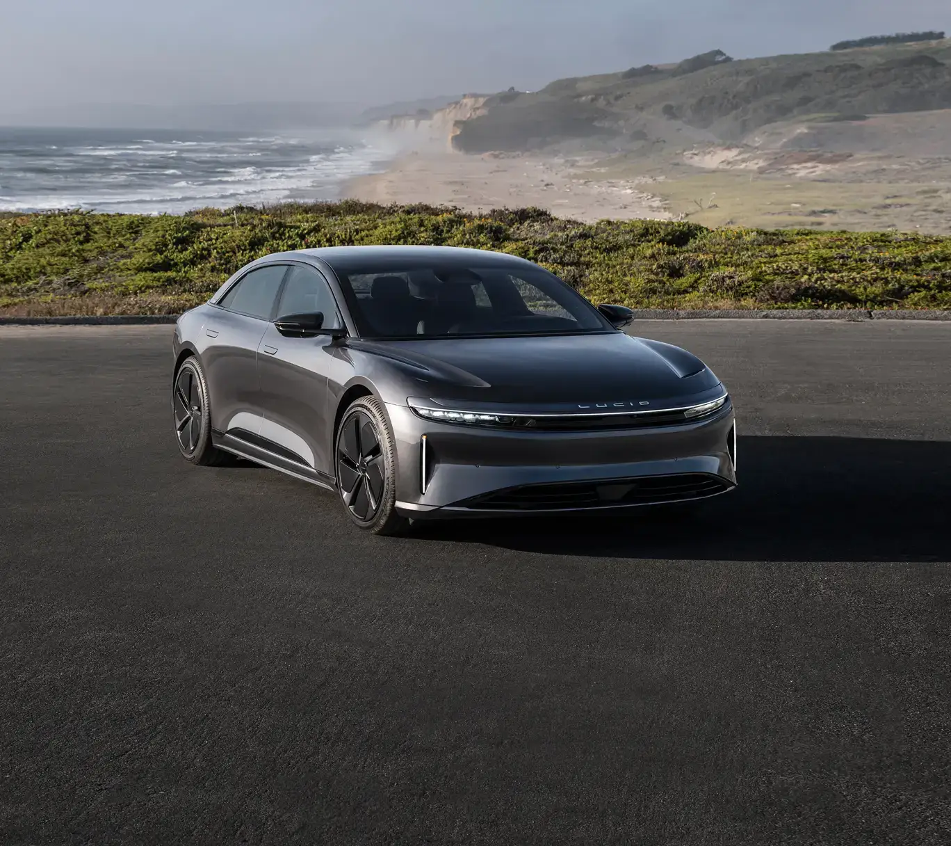 A sleek gray Lucid Air electric sedan parked on a coastal road with ocean, cliffs, and greenery in the background.