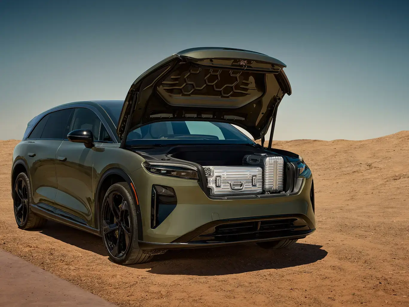 Green electric SUV with open front trunk displaying luggage storage capacity, parked in desert landscape under clear blue sky.