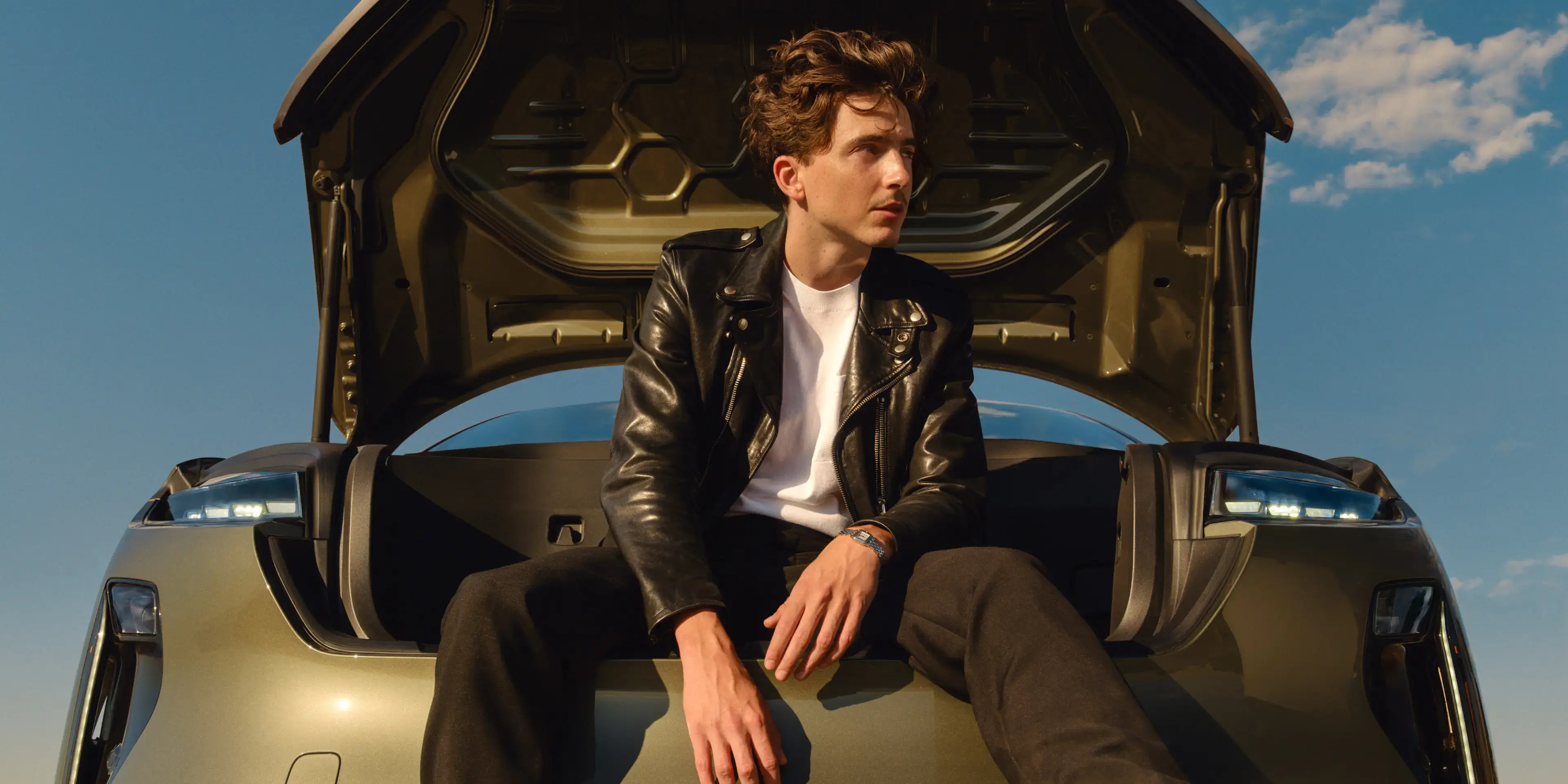 Timothée Chalamet in leather jacket sitting on open trunk of Lucid Gravity against blue sky with scattered clouds