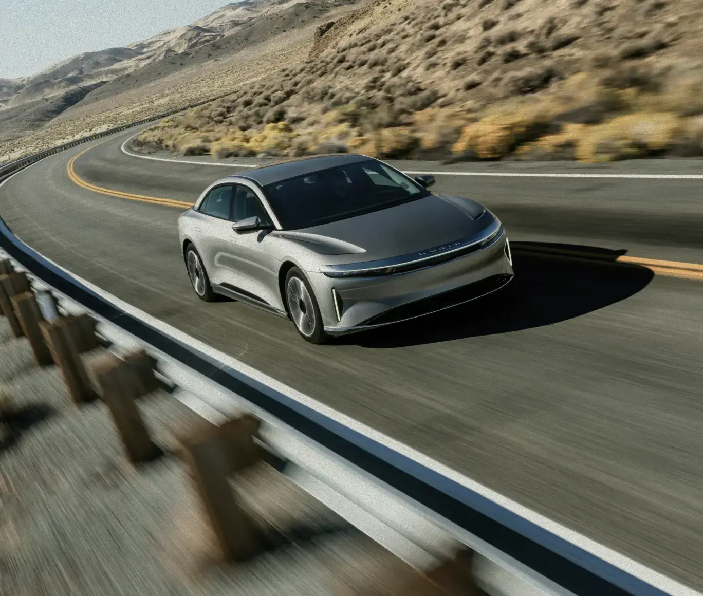 A silver Lucid Air electric sedan driving on a winding desert highway with rocky hills in the background.
