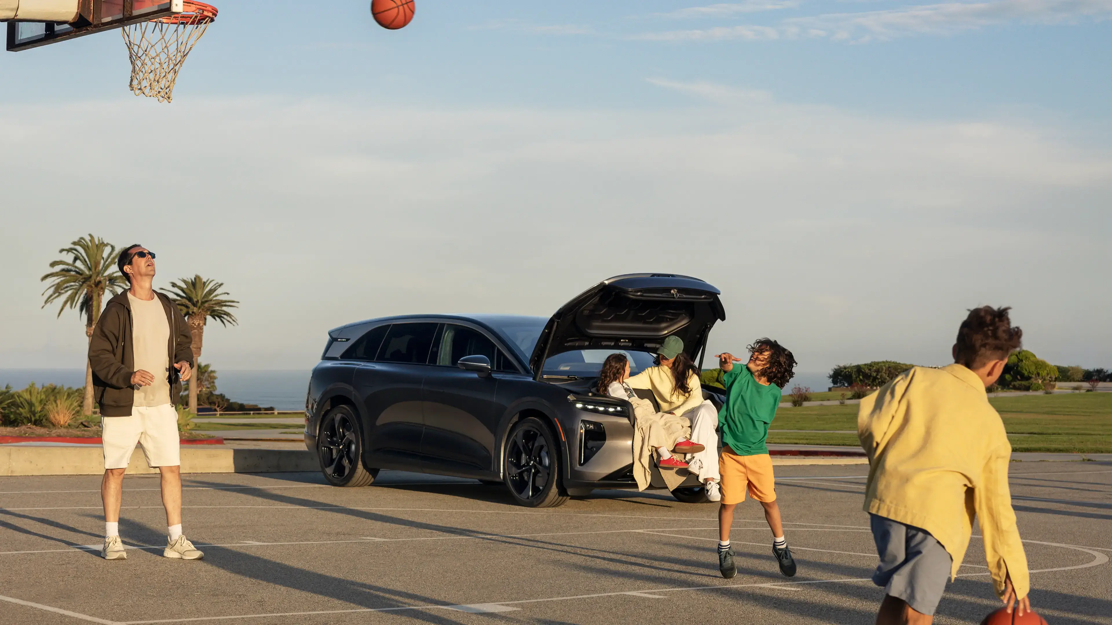 A child tosses a basketball in a parking lot beside a Lucid Gravity SUV with open frunk, where another person sits relaxing outdoors.