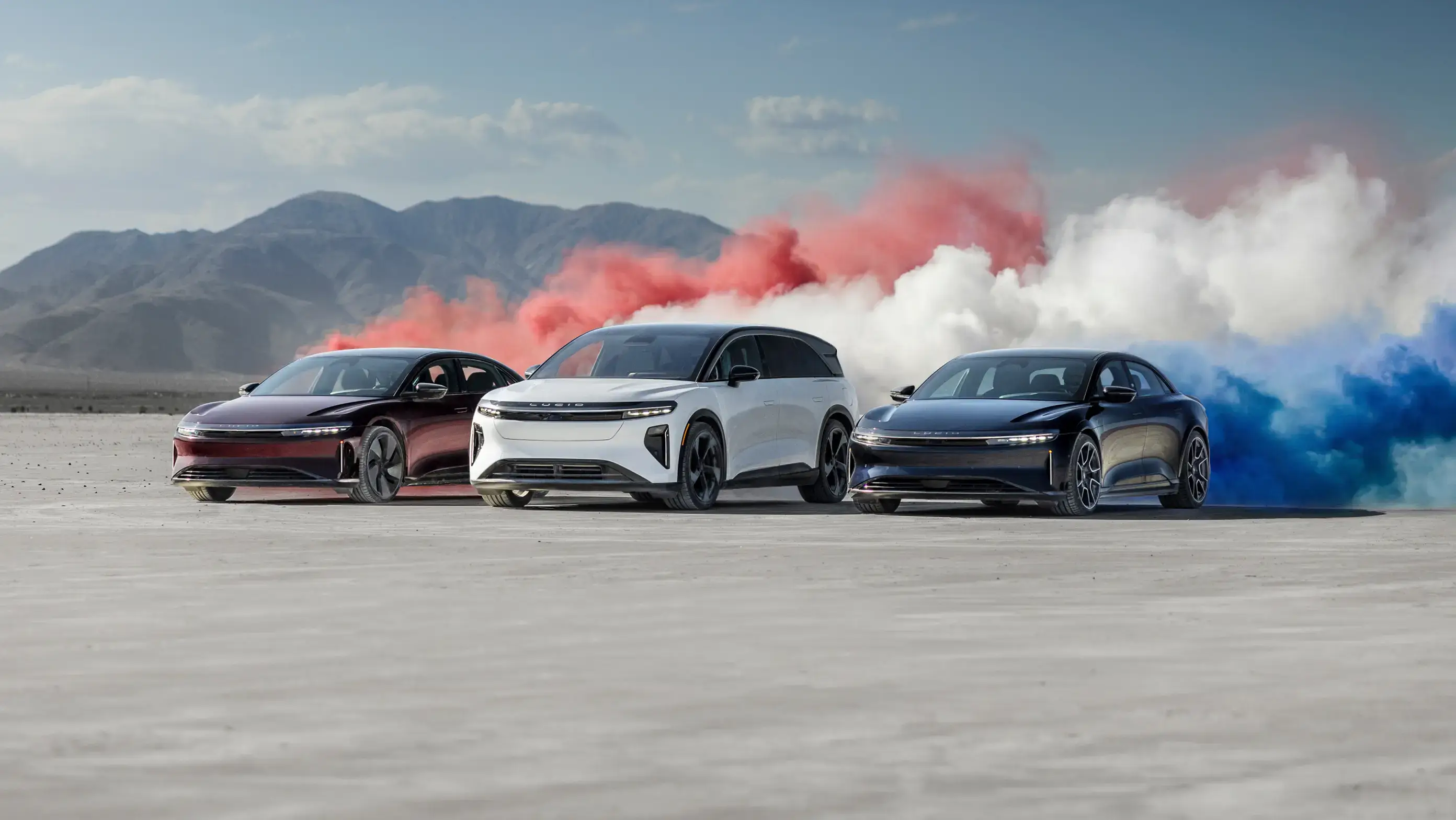 Three Lucid electric vehicles in red, white, and blue with colorful smoke clouds on a desert salt flat.